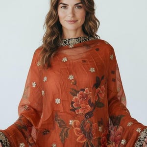May include: Rust-colored sheer fabric with floral embroidery and a black and gold border. The garment features large, detailed floral patterns in shades of red and orange. Small gold floral accents are scattered across the fabric.