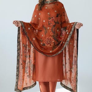 May include: A rust-colored salwar kameez ensemble. The outfit includes a long tunic, matching pants, and a sheer dupatta. The dupatta features a floral print with red and white flowers and a black and gold border. The outfit is worn with sandals.