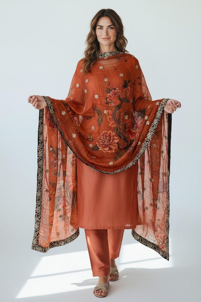 May include: A rust-colored salwar kameez ensemble. The outfit includes a long tunic, matching pants, and a sheer dupatta. The dupatta features a floral print with red and white flowers and a black and gold border. The outfit is worn with sandals.