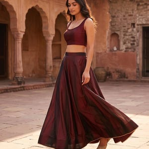 May include: A two-piece set consisting of a sleeveless, scoop-neck crop top and a floor-length, high-waisted skirt in a deep burgundy color. The skirt features a full, flowing silhouette with a subtle sheen and a thin gold trim along the hem.
