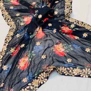 May include: A black sheer scarf with a floral pattern in red, blue, and yellow. The scarf has a gold embroidered border and scattered gold flower accents. The fabric is draped, showcasing the design.