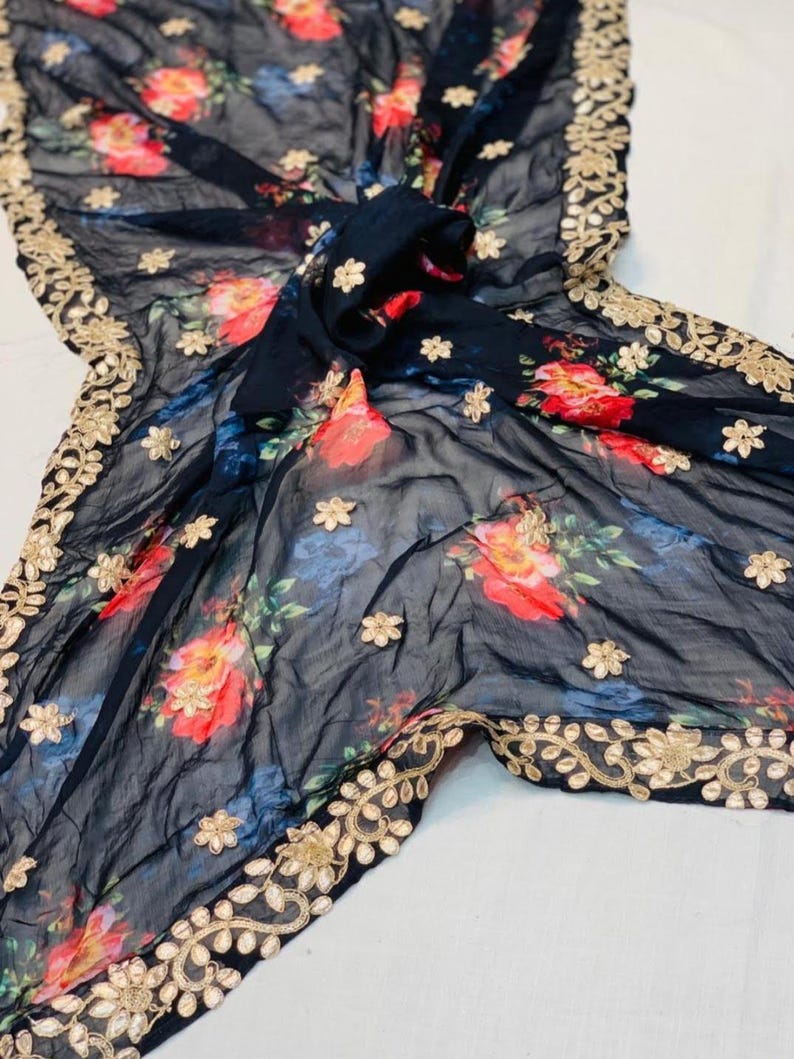 May include: A black sheer scarf with a floral pattern in red, blue, and yellow. The scarf has a gold embroidered border and scattered gold flower accents. The fabric is draped, showcasing the design.