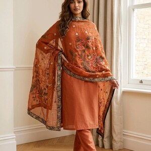May include: An orange salwar kameez ensemble featuring a long-sleeved tunic and matching pants. The sheer dupatta is adorned with floral patterns and a black and gold border. The outfit is worn with sandals.
