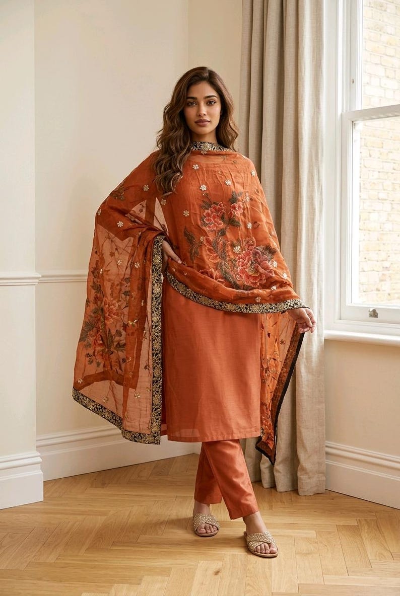 May include: An orange salwar kameez ensemble featuring a long-sleeved tunic and matching pants. The sheer dupatta is adorned with floral patterns and a black and gold border. The outfit is worn with sandals.