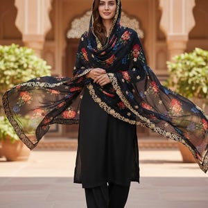 May include: A woman wearing a black salwar kameez with a floral print dupatta. The dupatta has a gold border and is draped over the head and shoulders. The outfit is worn with matching black pants and sandals.