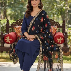 May include: A woman wearing a navy blue salwar kameez, a traditional South Asian outfit, sits on a wooden swing. The outfit includes a matching top and pants, and a black dupatta with floral embroidery. Gold-colored shoes complete the look.