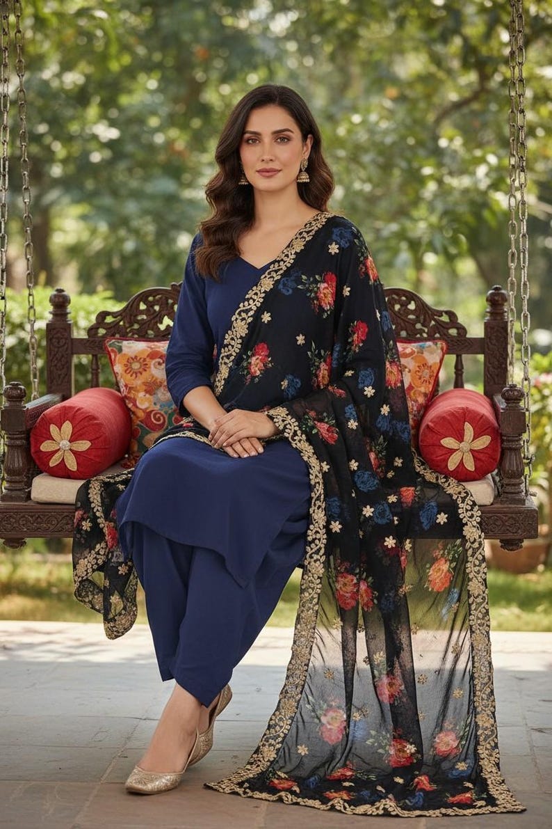 May include: A woman wearing a navy blue salwar kameez, a traditional South Asian outfit, sits on a wooden swing. The outfit includes a matching top and pants, and a black dupatta with floral embroidery. Gold-colored shoes complete the look.