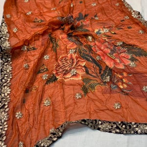 May include: An orange sheer scarf with a floral design in shades of red, orange, and green. The scarf is embellished with small, silver-colored sequins and has a black border with gold-colored accents.