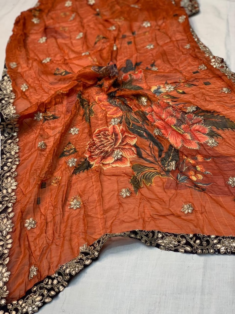 May include: An orange sheer scarf with a floral design in shades of red, orange, and green. The scarf is embellished with small, silver-colored sequins and has a black border with gold-colored accents.