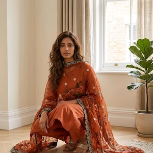 May include: Rust-colored salwar kameez set with floral embroidery. The ensemble includes a long-sleeved tunic, matching pants, and a sheer dupatta. The dupatta and neckline feature a black and gold border. The outfit is paired with gold sandals.