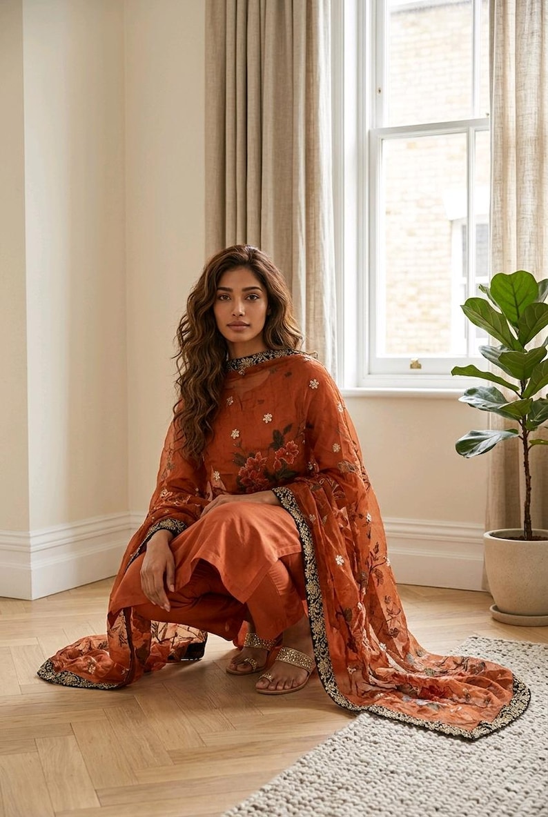 May include: Rust-colored salwar kameez set with floral embroidery. The ensemble includes a long-sleeved tunic, matching pants, and a sheer dupatta. The dupatta and neckline feature a black and gold border. The outfit is paired with gold sandals.