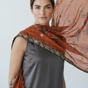 May include: A woman wearing a gray sleeveless dress and a sheer, rust-colored scarf. The scarf features floral patterns and gold embellishments along the edges. The scarf is draped over the shoulders and arms.