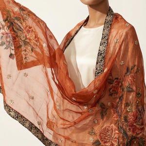 May include: A sheer, rust-colored scarf with floral embroidery and a black and gold border. The scarf features detailed embroidery of flowers in shades of red, orange, and green, with small, sparkling embellishments.