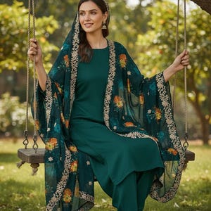 May include: A teal green salwar kameez ensemble. The outfit includes a tunic, pants, and a patterned shawl with floral and gold accents. The woman is sitting on a wooden swing outdoors.