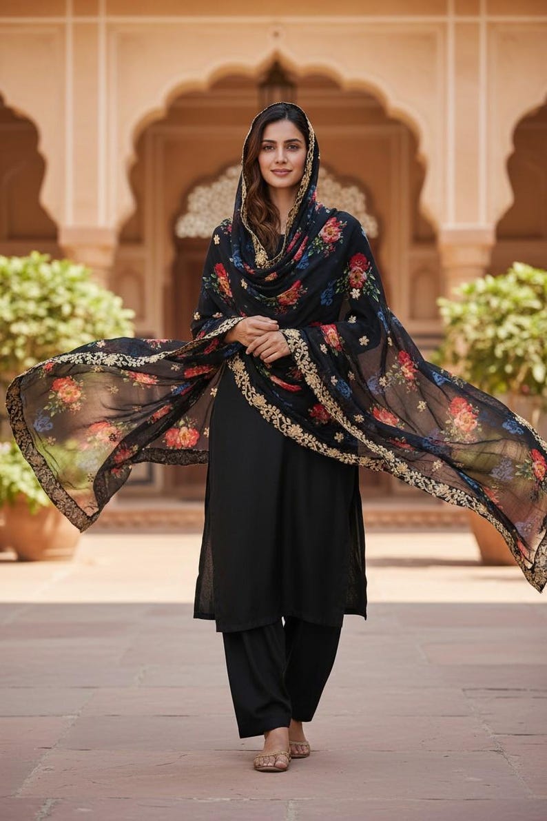 May include: A woman wearing a black salwar kameez with a floral print dupatta. The dupatta has a gold border and is draped over the head and shoulders. The outfit is worn with matching black pants and sandals.