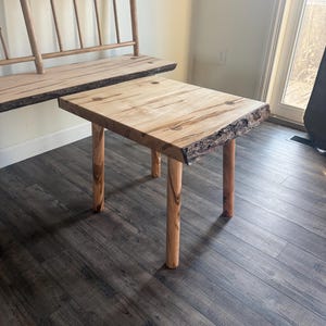 Könnte beinhalten: Kleiner, quadratischer Holztisch mit natürlicher Kante und vier runden Beinen. Die Tischplatte ist hell mit dunklen Maserungsmustern. Im Hintergrund ist eine passende Bank mit Rückenlehne zu sehen. Die Möbel sind aus Holz.