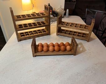 May include: Three wooden egg holders in various sizes. The largest has three tiers of egg storage, the medium has two, and the smallest holds a dozen eggs. The wood has a warm, brown stain, and the eggs are light brown.