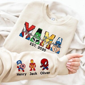 A person is holding a white t-shirt with a design featuring the Marvel superheroes Spider-Man, Captain America, and Iron Man.