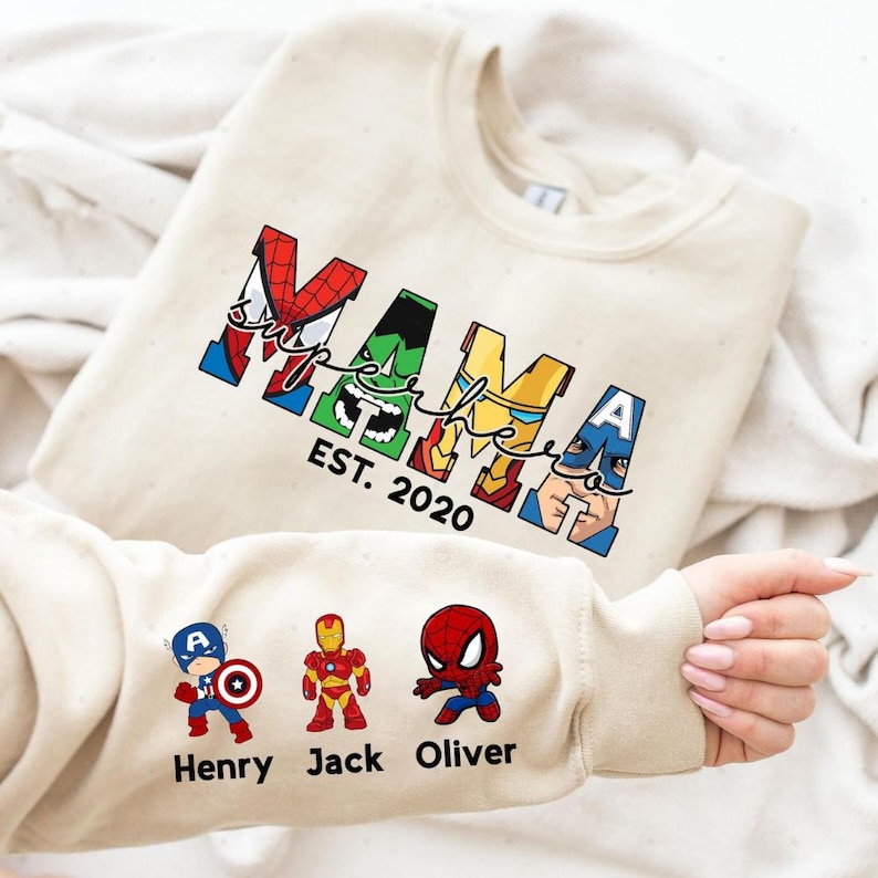 A person is holding a white t-shirt with a design featuring the Marvel superheroes Spider-Man, Captain America, and Iron Man.