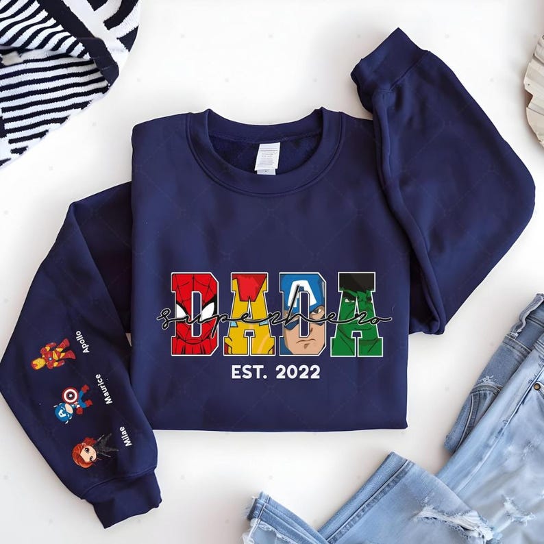 A navy blue sweatshirt with the word "Dada" printed on it, along with various superhero characters. The sweatshirt is laid out on a white surface, with a pair of jeans and a striped towel visible in the background.