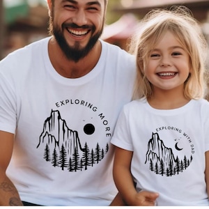 May include: White t-shirts with matching mountain and forest graphic designs. The adult shirt says "Exploring More" and the child's shirt says "Exploring with Dad". The design includes a mountain, trees, a sun, and a crescent moon.