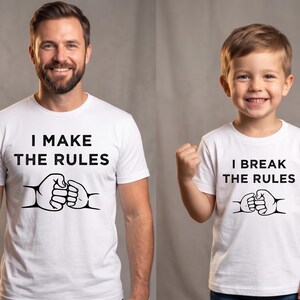 May include: Two white t-shirts with the text "I MAKE THE RULES" and "I BREAK THE RULES" in black. Each shirt features a graphic of two fists bumping. The shirts are worn by a man and a young child.