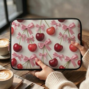 Könnte beinhalten: Eine schwarze Laptoptasche mit einem sich wiederholenden Muster aus roten Kirschen und rosa Schleifen auf weißem Hintergrund. Die Kirschen haben grüne Stiele und die Schleifen sind in der Mitte gebunden. Die Hülle hat einen schwarzen Rand.