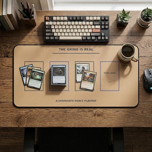 May include: A tan rectangular desk mat featuring the text THE GRIND IS REAL and A DESPERATE MAN'S PLAYMAT. The mat includes printed rectangular outlines for card placement, with several trading cards and a deck of cards arranged on its surface.