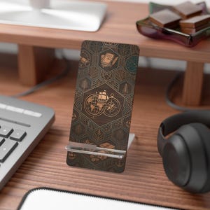 May include: Wooden phone stand with a travel-themed design. The stand features a dark brown background with a geometric pattern and engraved images of a globe, compass, ship, and other travel-related icons. The stand is designed to hold a phone upright.