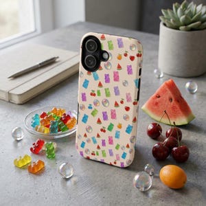 Op de afbeelding: Een beige smartphonehoesje met een herhalend patroon van kleurrijke gummibeertjes, watermeloenpartjes, kersen, sinaasappels en iriserende bubbels. Het hoesje heeft een zwarte camera-uitsparing en staat rechtop op een grijs oppervlak.