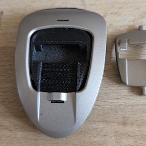 May include: A silver-colored electronic device with a black interior component and a separate silver-colored cover. The device is on a wooden surface. The device has a rectangular opening and a small, rectangular cover.