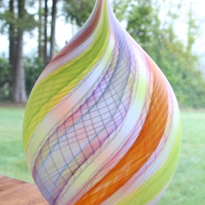 May include: A colorful, teardrop-shaped glass vase. The vase features a swirl design with bands of orange, yellow, green, purple, and white. The vase is sitting on a wooden surface, with a blurred green background.