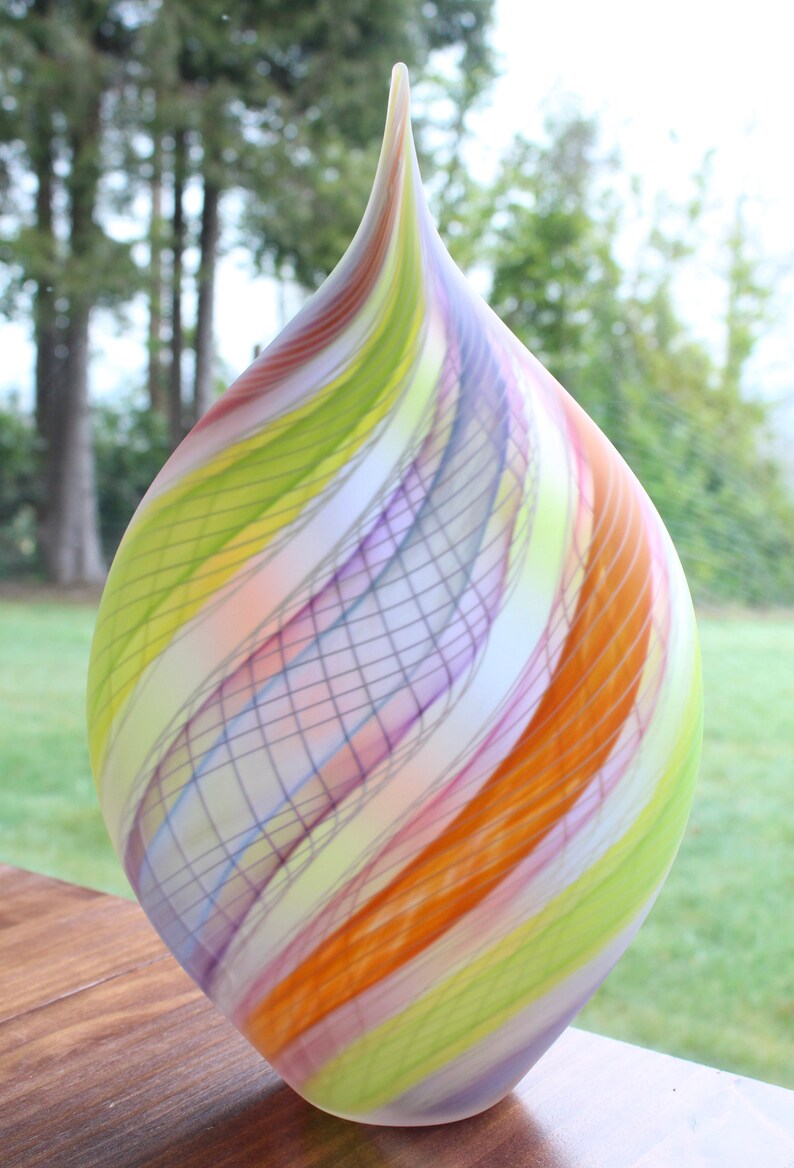 May include: A colorful, teardrop-shaped glass vase. The vase features a swirl design with bands of orange, yellow, green, purple, and white. The vase is sitting on a wooden surface, with a blurred green background.