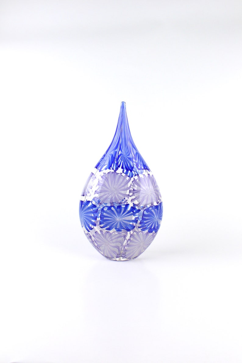 May include: A decorative glass vase in a teardrop shape. The vase is primarily blue with a pattern of white and blue floral designs. The vase has a long, pointed top and a rounded base. The glass appears to be hand-blown.