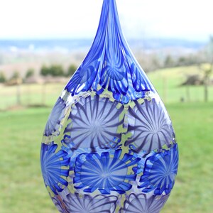 May include: A tall, teardrop-shaped glass vase with a pointed top. The vase features a pattern of blue and white floral designs. The vase is displayed on a wooden surface with a blurred green background.