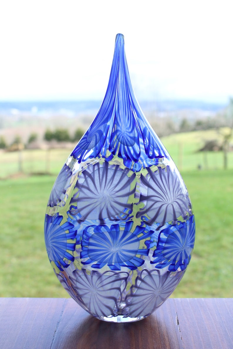 May include: A tall, teardrop-shaped glass vase with a pointed top. The vase features a pattern of blue and white floral designs. The vase is displayed on a wooden surface with a blurred green background.