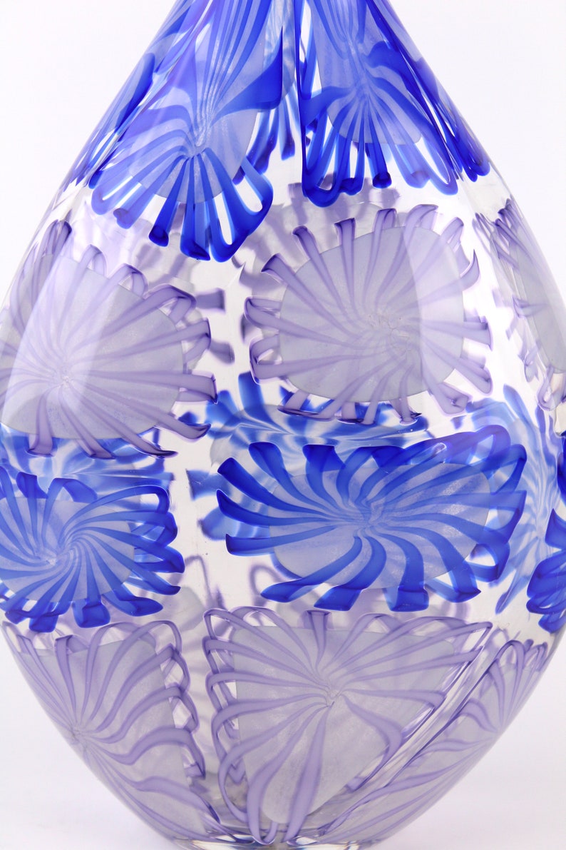 May include: A clear glass vase with a unique design. The vase features a pattern of swirling blue and lavender floral shapes. The vase has a rounded, elongated shape, and the design is visible throughout the glass.