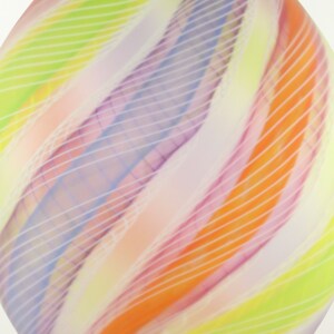 May include: Abstract glass art piece with a teardrop shape. The design features swirling bands of color, including orange, yellow, purple, and white, with thin white lines overlaying the colors. The overall effect is a soft, diffused appearance.