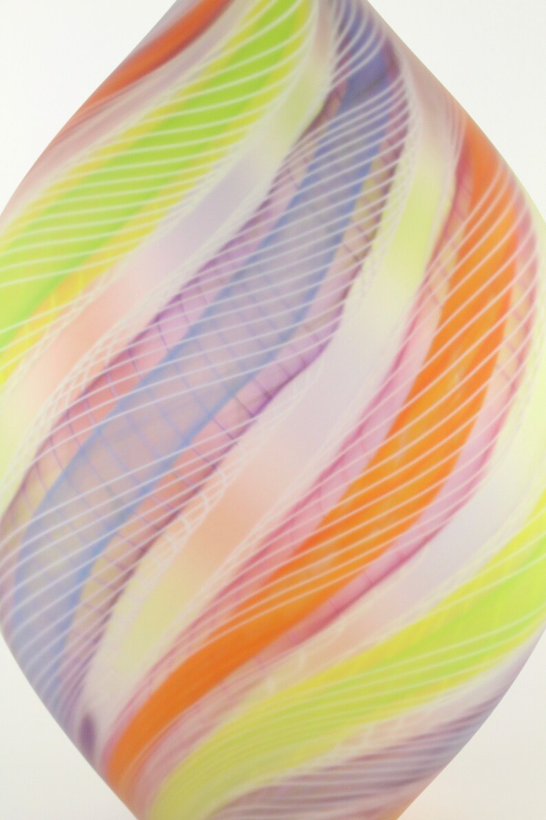 May include: Abstract glass art piece with a teardrop shape. The design features swirling bands of color, including orange, yellow, purple, and white, with thin white lines overlaying the colors. The overall effect is a soft, diffused appearance.