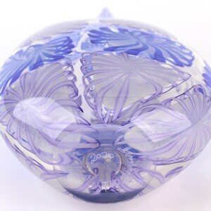 May include: A clear glass orb with a blue floral design. The orb has a rounded shape with a small, pointed top. The floral pattern is a series of swirling lines and shapes. The glass is transparent, allowing light to pass through.