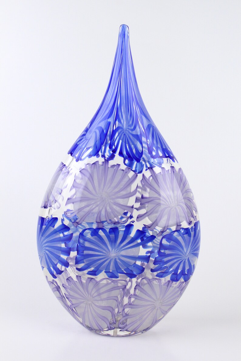 May include: A teardrop-shaped glass vase with a long, pointed top. The vase features a pattern of blue and lavender floral designs on a clear background. The top is a solid blue color.