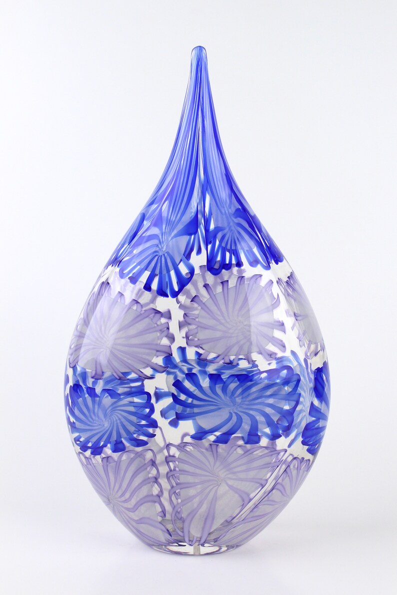 May include: A teardrop-shaped glass vase with a clear base and a pattern of blue and lavender swirls. The vase has a long, pointed top and is suitable for home decor.