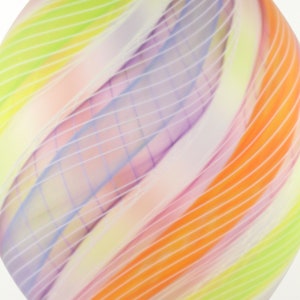May include: Abstract glass art piece with a diamond shape. The artwork features a blend of colors, including orange, yellow, green, and purple, with white linear accents. The design has a flowing, curved pattern.