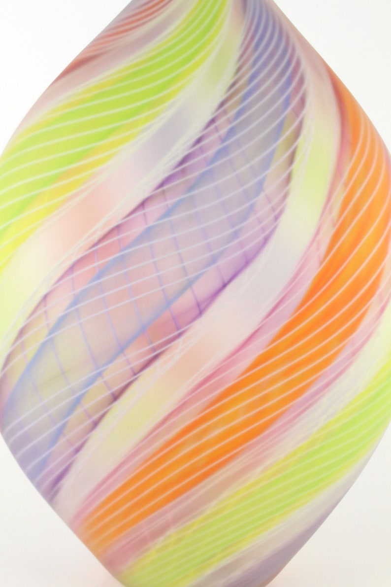 May include: Abstract glass art piece with a diamond shape. The artwork features a blend of colors, including orange, yellow, green, and purple, with white linear accents. The design has a flowing, curved pattern.