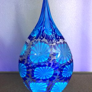 May include: A teardrop-shaped glass vase in shades of blue. The vase features a pattern of radiating blue circles and vertical blue stripes at the top. The vase is transparent, allowing light to pass through.