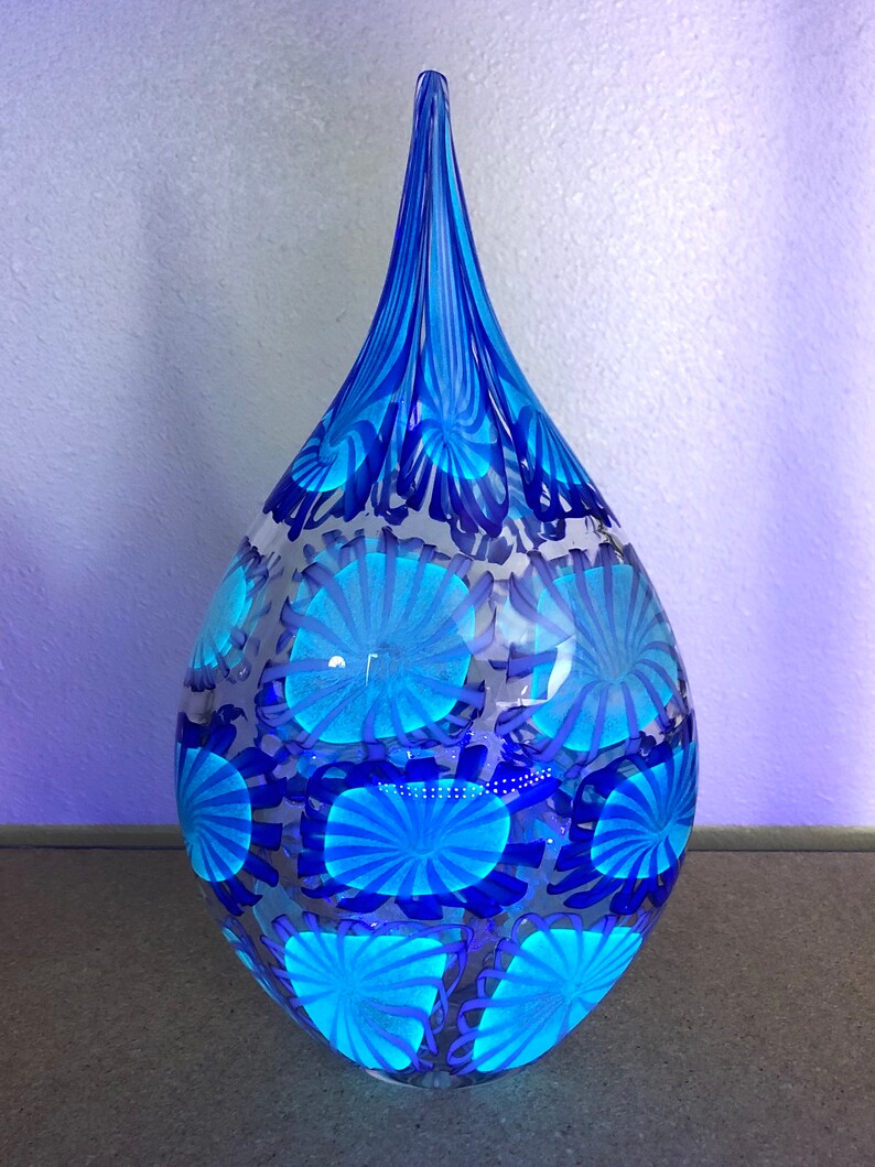 May include: A teardrop-shaped glass vase in shades of blue. The vase features a pattern of radiating blue circles and vertical blue stripes at the top. The vase is transparent, allowing light to pass through.