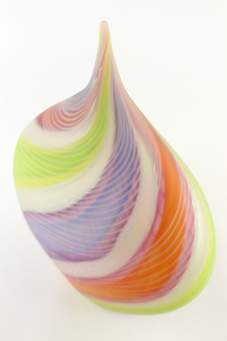 May include: A colorful, teardrop-shaped glass art piece. The sculpture features swirling bands of orange, pink, purple, green, and white. The glass has a smooth, glossy finish and a pointed top. The artwork is set against a white background.
