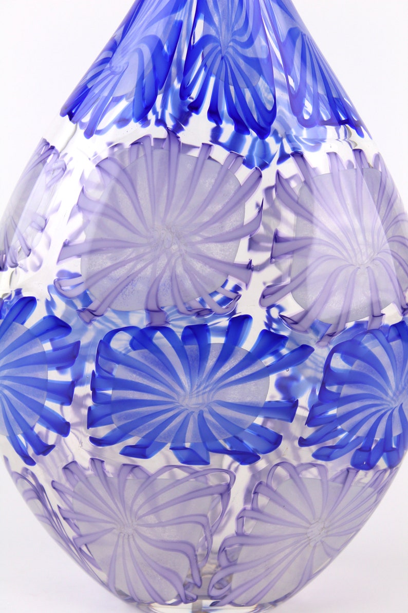 May include: A clear glass vase with a teardrop shape. The vase is decorated with a pattern of blue and lavender floral designs. The blue designs are more prominent near the top of the vase.