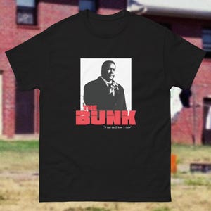 Könnte beinhalten: Ein schwarzes T-Shirt mit kurzen Ärmeln und einem schwarz-weißen Fotodruck einer Person im Anzug. Unter dem Bild steht der Text THE BUNK in großen roten Großbuchstaben, gefolgt von dem kleineren weißen Text A man must have a code.