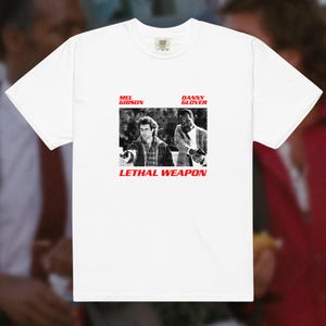 May include: A white short-sleeved t-shirt featuring a black and white graphic of Mel Gibson and Danny Glover holding handguns. The text "MEL GIBSON" and "DANNY GLOVER" appears in red above the image, with "LETHAL WEAPON" in red text below.